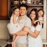 Satisfied man with daughter and woman with son embracing embracing each other while standing against wooden cupboard in kitchen and looking at camera