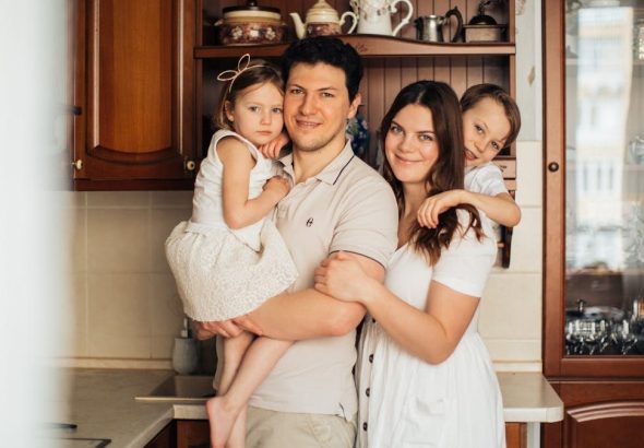 Satisfied man with daughter and woman with son embracing embracing each other while standing against wooden cupboard in kitchen and looking at camera