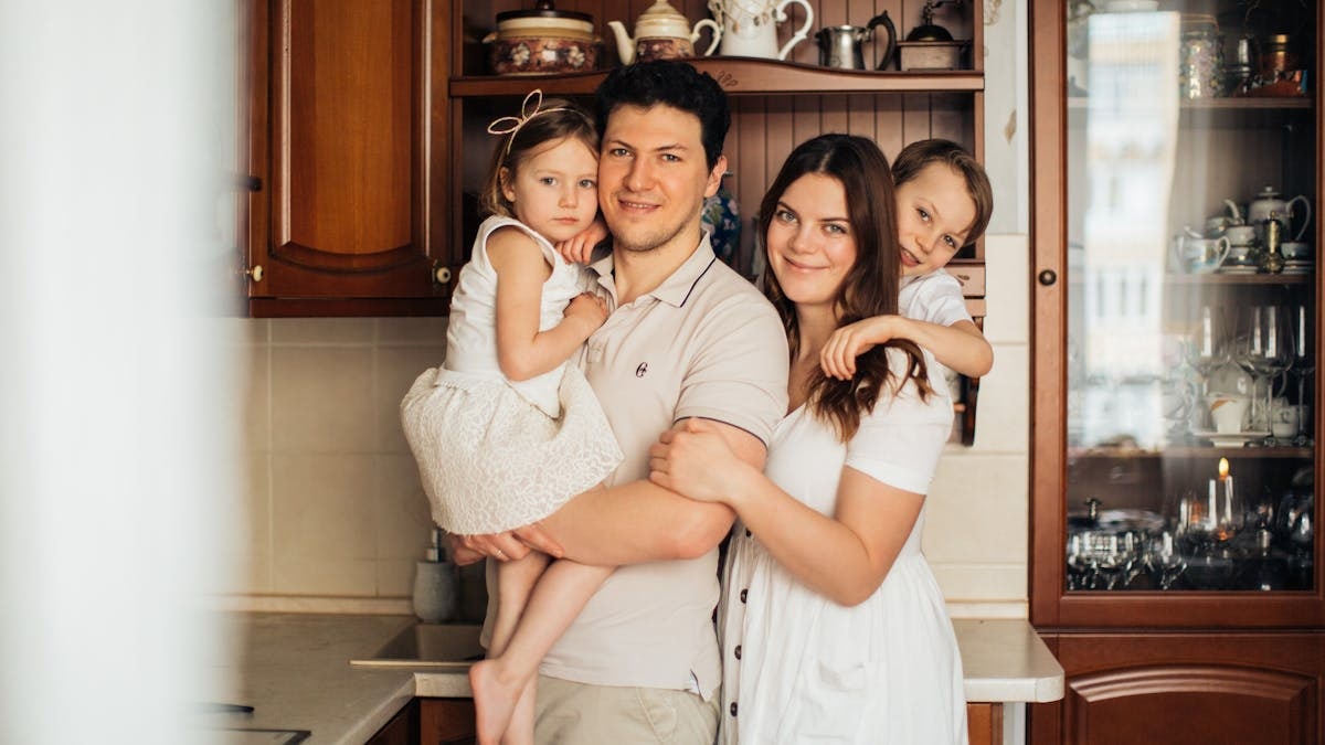 Satisfied man with daughter and woman with son embracing embracing each other while standing against wooden cupboard in kitchen and looking at camera
